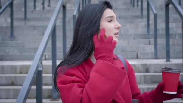 Close-up portrait of anxious Caucasian woman standing outdoors with coffee cup and talking on the phone. Young elegant girl in red coat waiting for meeting on city street. Lifestyle, break, meetings. — 图库视频影像