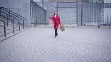 Portrait of young cheerful Caucasian woman walking to camera, winking, and leaving. Elegant stylish lady in red coat going home after shopping with bags. Style, fashion, shopaholism.