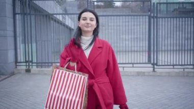 Middle shot front view of elegant young lady in red coat walking in city with shopping bags. Positive Caucasian woman looking at camera and smiling. Shopaholism, lifestyle, joy.