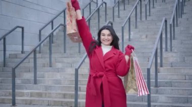 Portrait of cheerful Caucasian girl in red coat dancing with shopping bags outdoors. Excited female shopaholic satisfied with purchases. Joy, lifestyle, shopaholism.