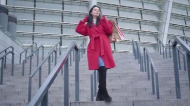 Bottom view of young stylish Caucasian woman standing on stairs outdoors and looking at camera. Portrait of elegant lady posing in city with shopping bags. Elegance, lifestyle, shopaholism.