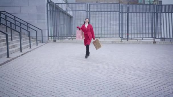 Retrato de una joven y alegre mujer caucásica caminando hacia la cámara, guiñando el ojo y saliendo. Elegante y elegante dama de abrigo rojo que vuelve a casa después de ir de compras con bolsas. Estilo, moda, compras . — Vídeo de stock