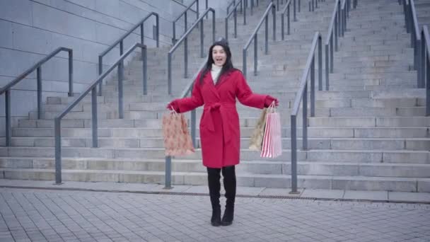 Largo tiro de mujer caucásica emocionada bailando con bolsas de compras en el fondo de la escalera en la ciudad. Chica joven de moda en abrigo rojo divertirse después de ir de compras. Estilo de vida, disfrute, consumismo . — Vídeo de stock