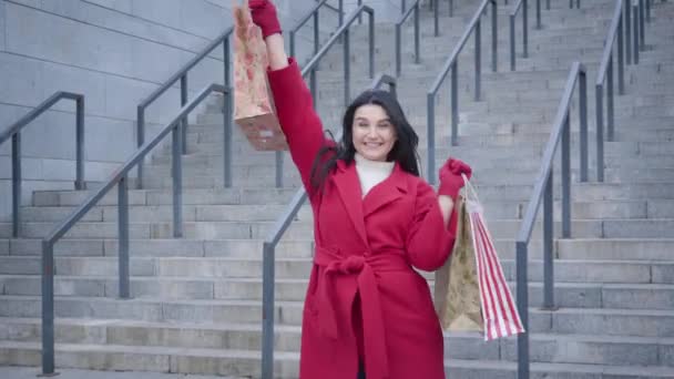 Portrait of cheerful Caucasian girl in red coat dancing with shopping bags outdoors. Excited female shopaholic satisfied with purchases. Joy, lifestyle, shopaholism. — Stockvideo