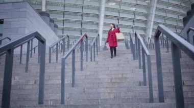 Long shot of young smiling Caucasian lady in red coat and black boots walking down the stairs to camera outdoors. Cheerful female shopaholic walking in city after shopping. Consumerism, lifestyle.