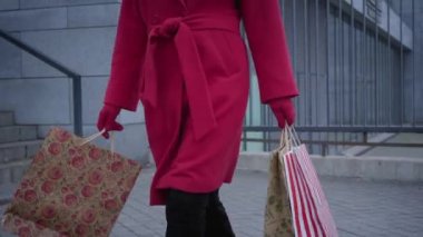 Unrecognizable elegant lady in red coat, gloves, and black boots walking with shopping bags in city. Stylish young woman shopping outdoors. Shopaholism, consumerism, lifestyle, fashion.