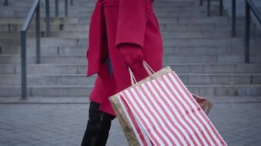 Side view of unrecognizable woman in red coat and gloves walking with shopping bags in city. Young fashionable lady strolling at the background of stairs outdoors. Fashion, lifestyle, consumerism.