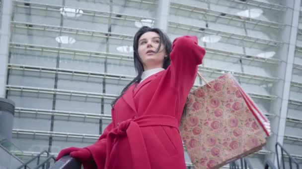 Middle shot bottom view of elegant Caucasian brunette girl in red coat standing outdoors with shopping bags. Young beautiful lady with black hair looking around in city. Lifestyle, shopaholism. — Stock Video