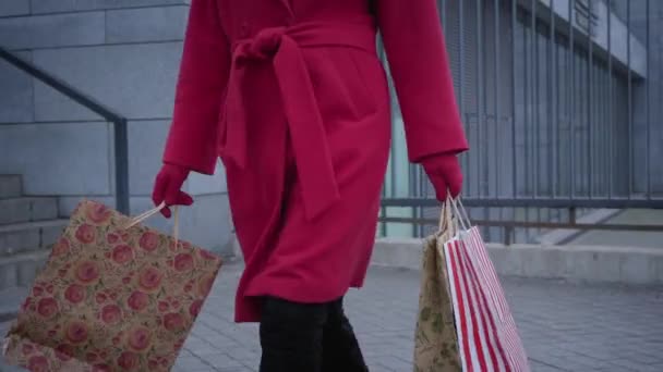 Невпізнавана елегантна леді в червоному пальто, рукавичках і чорних черевиках, що ходять з сумками в місті. Стильна молода жінка купує на відкритому повітрі. Шопоголізм, споживання, спосіб життя, мода . — стокове відео