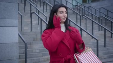 Young cheerful Caucasian girl talking on the phone and smiling standing on city street. Cheerful satisfied woman in red coat holding shopping bags and jumping up. Style, fashion, shopaholism.