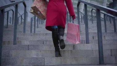 Camera following unrecognizable stylish lady in red coat and black boots walking up the stairs in city with shopping bags. Young stylish lady with black hair doing shopping outdoors.