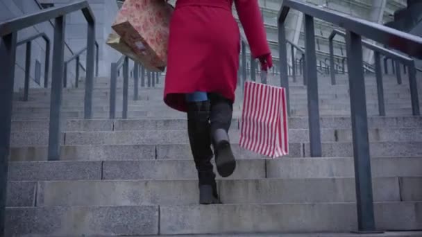 Kamera sledující nepoznatelnou stylovou dámu v červeném kabátě a černých botách kráčející po schodech ve městě s nákupními taškami. Mladý stylový dáma s černými vlasy dělat nákupy venku. — Stock video