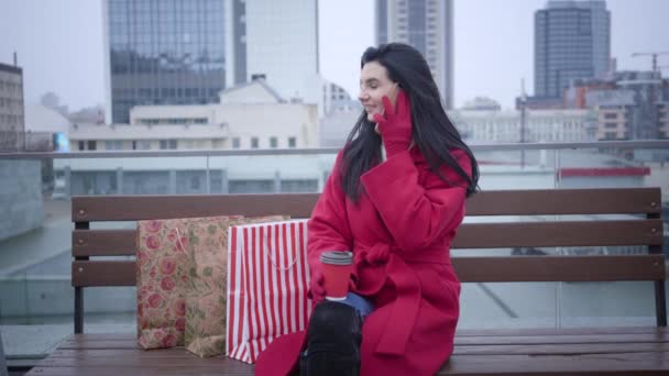 Portrait of positive cute Caucasian woman in red coat sitting on bench in the city with shopping bags and talking on the phone. Young lady enjoying rainy day outdoors. Lifestyle, joy, shopaholism. — Stockvideo