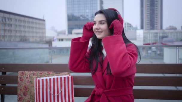 Joven y alegre mujer caucásica poniéndose auriculares y escuchando música en la ciudad. Chica positiva en abrigo rojo descansando después de comprar sentado en el banco al aire libre. Estilo de vida, compras, consumismo . — Vídeo de stock