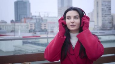 Portrait of confident Caucasian girl in red coat and gloves putting on headphones as sitting in city under light rain or snow. Positive girl listening to music in earphones. Hobby, lifestyle, leisure.