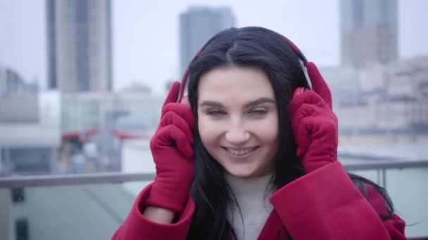 Close-up face of smiling young Caucasian girl with black hair and green eyes putting on earphones in city. Cheerful cute woman listening to music outdoors. Lifestyle, beauty, hobby. — Stockvideo