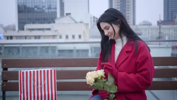 Retrato de una hermosa joven caucásica sentada en el banco de la ciudad con ramo de flores y bolsa de compras. Feliz señora con estilo oliendo rosas amarillas y sonriendo. Romance, estilo de vida, felicidad . — Vídeo de stock