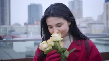 Close-up portrait of young Caucasian woman with black hair smelling yellow roses and smiling. Charming lady standing outdoors with a bouquet of flowers. Joy, happiness, lifestyle.
