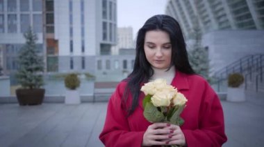 Portrait of romantic young Caucasian woman smelling yellow roses and looking at camera. Fashionable lady with black hair dressed in red coat posing in city in autumn day. Joy, happiness, lifestyle.