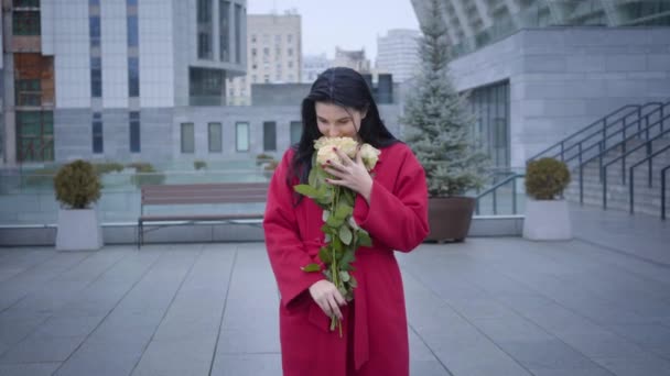 Camera approaching to happy young lady smelling yellow roses under light rain in city. Beautiful Caucasian woman holding bouquet of flowers and smiling. Joy, happiness, lifestyle. — Stockvideo