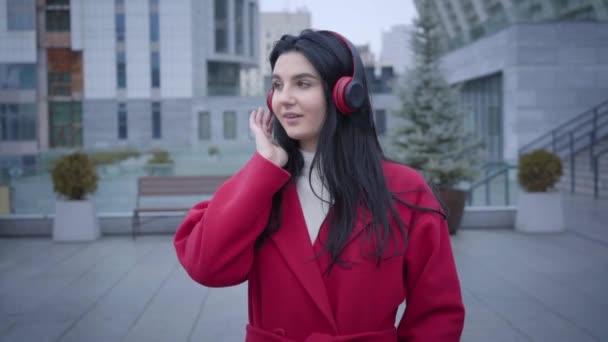 Портрет молодої веселої кавказької жінки, що танцює в навушниках на вулицях міста. Дівчина в червоному пальто слухає музику в навушниках. Спосіб життя, хобі, радість. — стокове відео