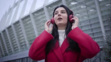 Bottom view of joyful Caucasian girl listening to music in headphones and dancing. Positive attractive woman enjoying autumn day in city. Fun, joy, happiness, lifestyle.