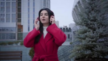 Portrait of cheerful cute girl in red coat walking and dancing as listening to music in earphones. Positive Caucasian stylish lady strolling in city in autumn day. Fun, leisure, hobby, lifestyle.