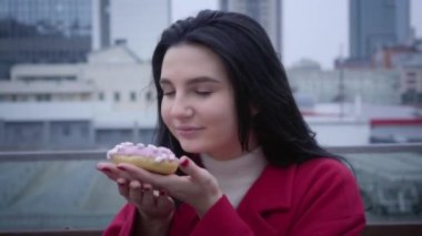Close-up portrait of young smiling Caucasian woman smelling donut and looking at camera. Cheerful woman having break in city under light rain. Lifestyle, enjoyment, happiness.
