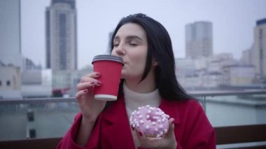 Close-up of cheerful Caucasian girl with green eyes and black hair drinking coffee and eating sweet tasty donut outdoors. Young confident woman resting in city and smiling. Fun, joy, lifestyle, break.