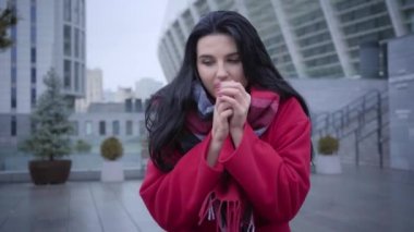 Freezed Caucasian woman blowing on hands and rubbing body. Beautiful brunette girl standing on city street in autumn red coat and warm scarf. Fashion, lifestyle.