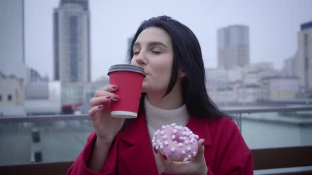 Close-up van vrolijke blanke meisje met groene ogen en zwart haar drinken van koffie en het eten van zoete smakelijke donut buiten. Jonge zelfverzekerde vrouw rustend in de stad en lachend. Plezier, vreugde, levensstijl, pauze. — Stockvideo