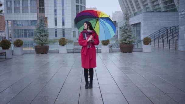 Довгий постріл позитивної молодої кавказької жінки з багатокольоровою парасолькою, що обертається по міській вулиці. Весела дівчина в стильному червоному пальто посміхається до дощового осіннього дня. Спосіб життя, щастя, радість. — стокове відео