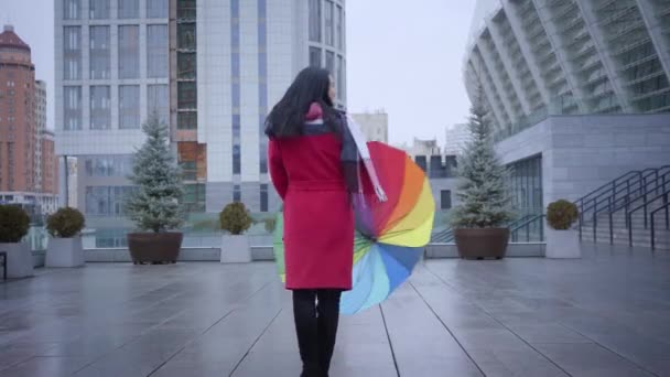 Kamera se blíží k pozitivní mladé ženě, která se otáčí s barevným deštníkem. Roztomilá veselá běloška s černými vlasy v červeném kabátě si užívá deštivého podzimního dne ve městě. Zábava, životní styl, radost. — Stock video