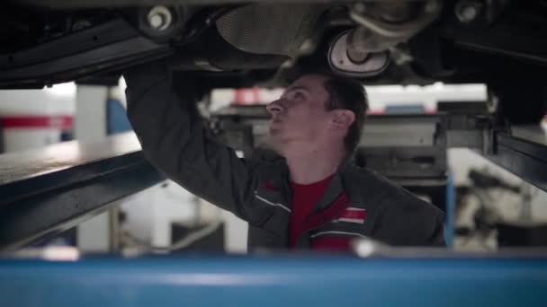 Retrato de un trabajador caucásico serio en uniforme mirando la parte inferior del coche en el taller de reparación. Ingeniero de mantenimiento masculino seguro que arregla la rotura del automóvil. Servicio, seguros, industria automotriz . — Vídeo de stock