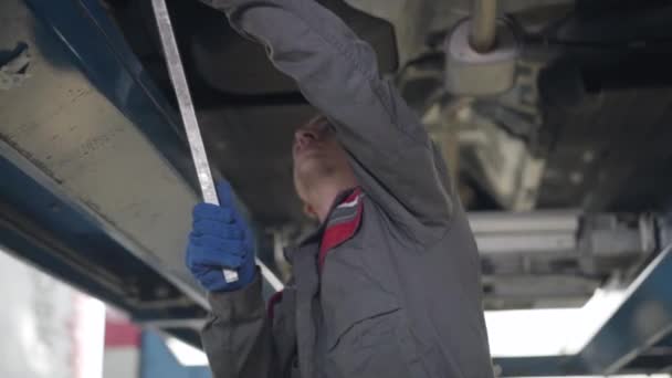 Young professional male maintenance engineer repairing car breakage. Confident man using equipment to fix automobile in repair shop. Service, tuning, replacement, industry. Camera moving upwards. — Stockvideo