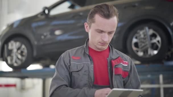 Close-up of professional male maintenance engineer standing in repair shop and using tablet. Caucasian auto mechanic thinking on fixing of broken car at the background. Service, industry, tuning. — 图库视频影像