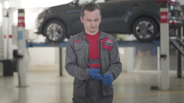 Střední záběr pozitivního mladého bělošského automechanika, jak si sundává ochranné rukavice, dívá se do kamery a ukazuje palec nahoru. Muž v pracovním oděvu pózuje v opravně. Automobilový průmysl, pojištění automobilů, ladění. — Stock video