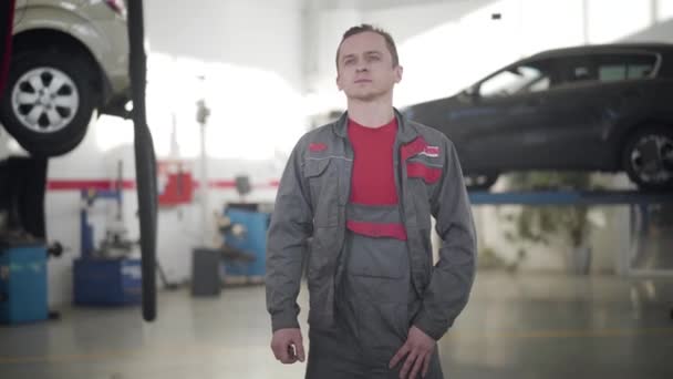 Plano medio del hombre caucásico reflexivo en ropa de trabajo mirando alrededor de coches rotos en el taller de reparación. Profesional mecánico de automóviles masculino en el trabajo. Profesión, estilo de vida, industria automotriz . — Vídeos de Stock