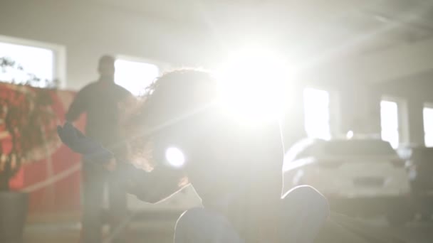 Hermosa joven mujer caucásica sentada en los rayos del sol en el taller de reparación de automóviles y sonriendo. Precioso mecánico de automóviles femenino con el pelo rizado morena posando a la luz del sol en el lugar de trabajo. Lento. . — Vídeo de stock