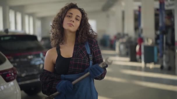 Slowmo portrait of young sensual Caucasian brunette woman with curly hair standing in sunrays at workplace and looking at camera. Female auto mechanic posing in car repair shop. Slow motion. — Stock Video
