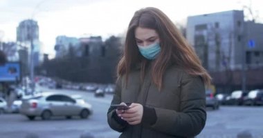 Şehir sokaklarında koruyucu maskeli ve akıllı telefon kullanan endişeli bir kadının portresi. Genç kız Coronavirus 'la ilgili haberleri okumakla meşgul. Covid-19, salgın, tehlike. Sinema 4k Prores Merkezi.