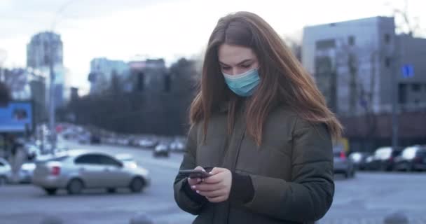 도시 거리에 서서 보호용 마스크를 쓰고 스마트폰을 사용하는 걱정하는 여성의 사진. 바쁜 소녀가 코로나 바이러스에 대한 뉴스를 읽고 있습니다. Covid-19, 세계적 유행병, 위험. 영화 4K 전립선 HQ. — 비디오