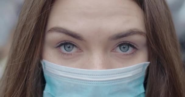 Close-up de olhos cinzentos de mulher jovem em máscara protetora olhando para a câmera. Retrato de morena linda menina ao ar livre. Epidemia, cuidados médicos, Covid-19. Cinema 4k ProRes HQ . — Vídeo de Stock