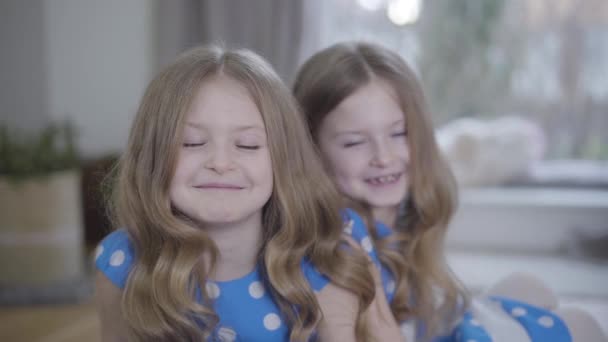 Portrait de jolie petite fille brune caucasienne aux yeux gris regardant la caméra et souriant. Sa sœur jumelle assise à l'arrière-plan et parlant avec sa sœur. Joie, bonheur, unité, style de vie . — Video