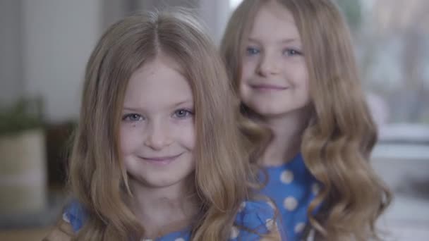 Zwei kleine hübsche kaukasische Zwillinge, die sich anschauen und lächelnd in die Kamera schauen. glückliche süße Kinder posieren zu Hause. Lebensstil, Glück, Freizeit, Einheit. Kamera entfernt sich. — Stockvideo