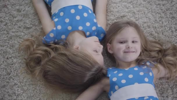 Вид сверху на двух симпатичных кавказских сестер-близнецов, лежащих на мягком ковре и улыбающихся. Камера двигается по лицам симпатичных брюнеток, отдыхающих дома. Досуг, образ жизни, единство . — стоковое видео