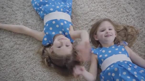Top view of cute little Caucasian girls waving at camera and smiling. Joyful brunette twin sisters having fun as resting at home. Lifestyle, family, unity, leisure. — Stock Video