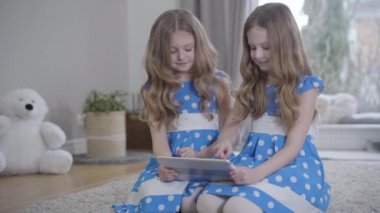 Evde tablet kullanan benzer elbiseli ikiz kız kardeşlerin portresi. Beyaz tenli esmer kızlar yumuşak halıda oturuyorlar. Yaşam tarzı, aile, eğlence..