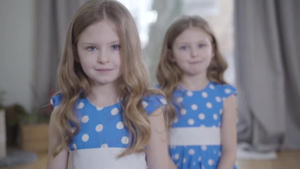 Caméra approchant de deux belles petites filles caucasiennes posant à l'intérieur. Brunette jumelles dans des robes similaires regardant la caméra et souriant. Concentré sur le visage de l'enfant au premier plan . — Video