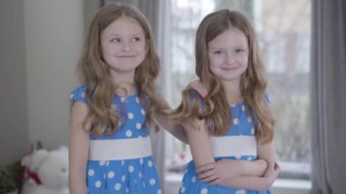Gülümseyen esmer beyaz ikiz kız kardeşler benzer noktalı mavi elbiseler giyip evde poz veriyorlar. İçerideki kameraya bakan pozitif küçük kız kardeşler. Neşe, mutluluk, birlik, rahatlık.